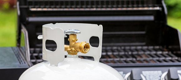 A white propane tank with a gold spout and silver knob is in sharp focus in the foreground, positioned in front of a black grill with silver accents. The grill, slightly blurred in the background, features two layers of grates.